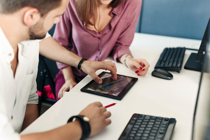 A man is showing a woman something on a tablet. Both are seated along a table that has computer keyboards on it.