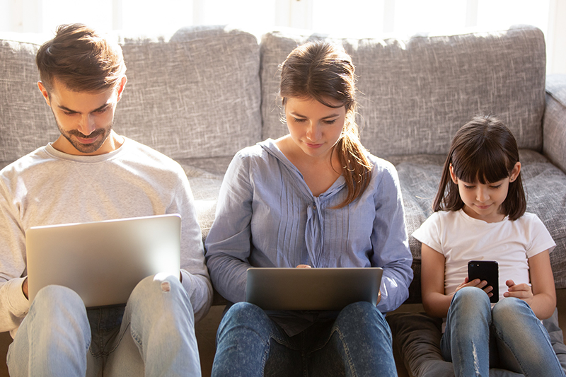 Three people sitting side by side in front of a couch. One is on a laptop, one is on a tablet, and one is on a smartphone.