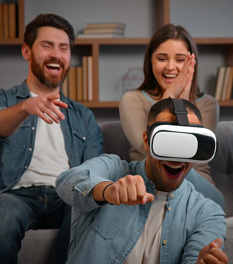 Photograph of man playing a video game wearing a virtual reality headset. Friends are laughing behind him.